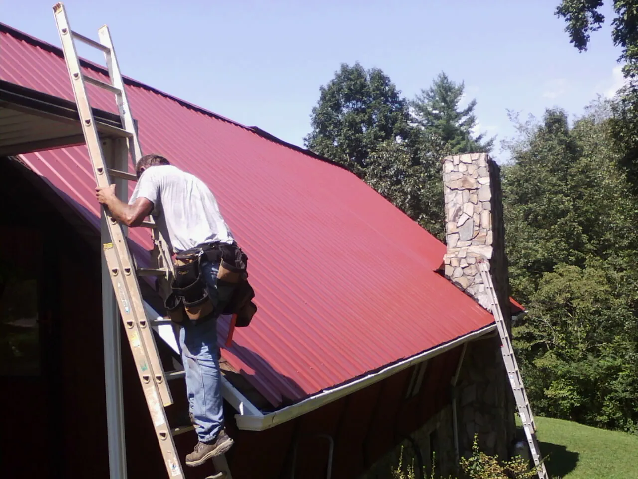 Roofer on a ladder beside a red metal roof and stone chimney — Soffit & Fascia Repair by Roofing Brothers in Rumford