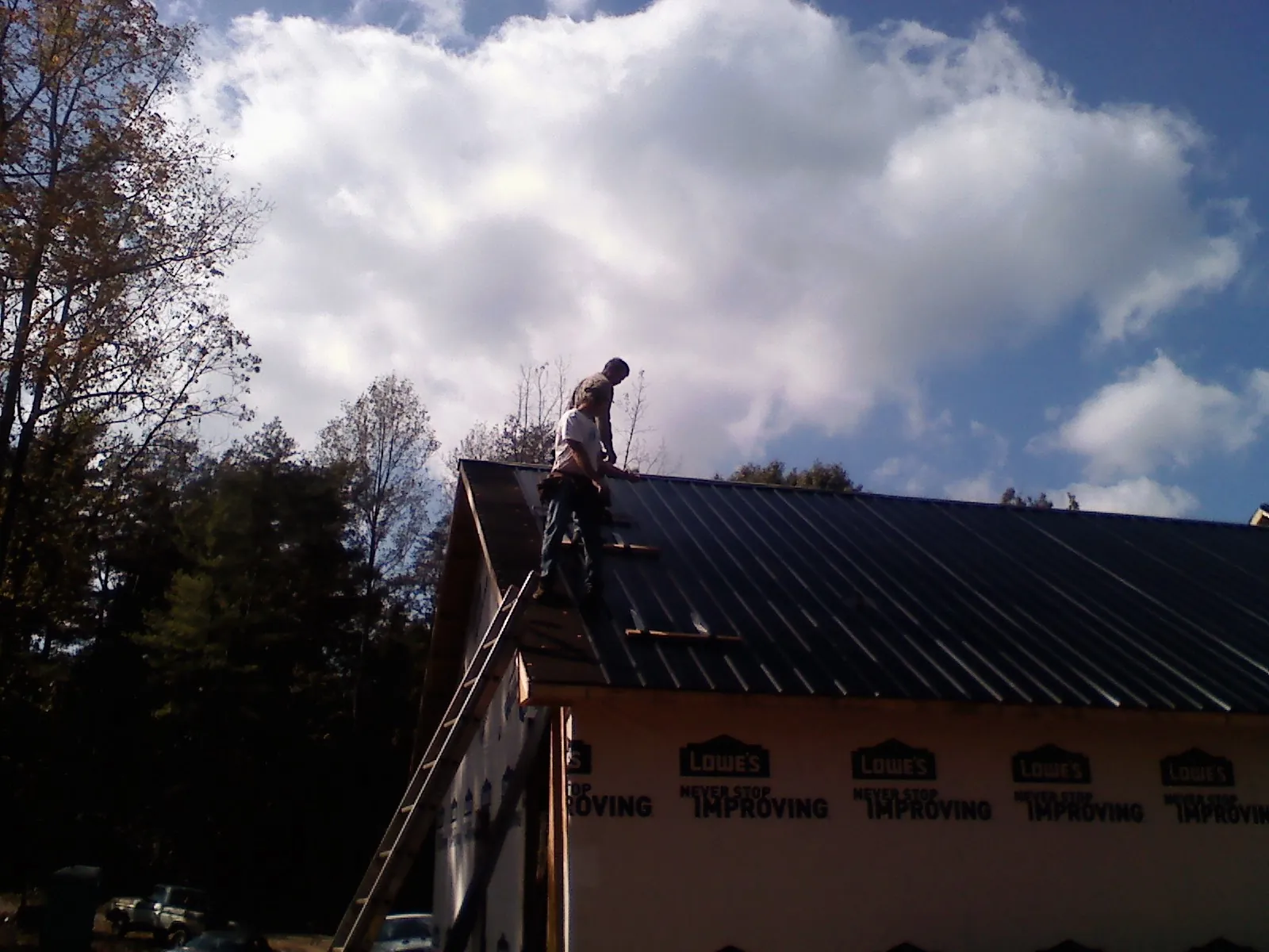 Roofer fastening dark metal panels high against a cloudy sky, from a Roof Inspection job in Rumford